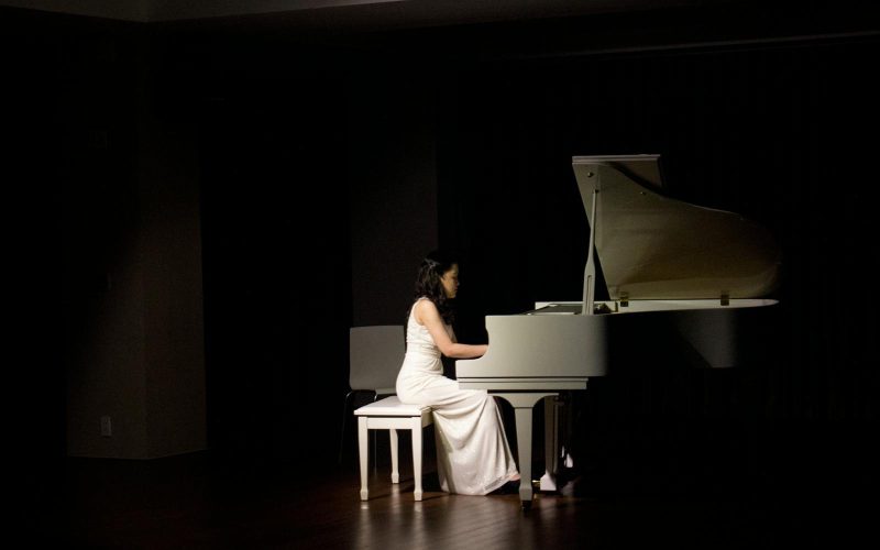A woman in a white dress plays a grand piano under stage lights, creating a dramatic performance ambiance.